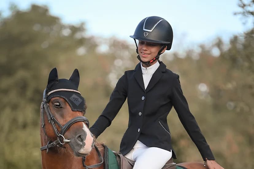 Jeune cavalière en selle sur un poney bai, bombe et veste noires, fin de journée